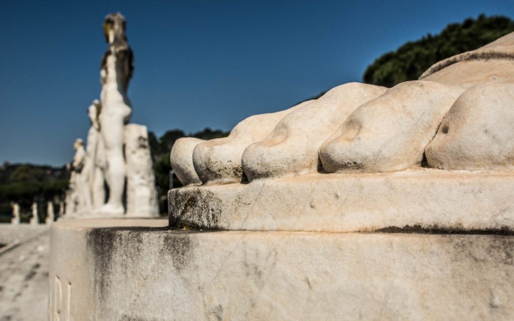 Zbliżenie na stopy olbrzymiego, marmurowego posągu sportowca na tle rzędu podobnych rzeźb na rzymskim Stadionie Marmurów