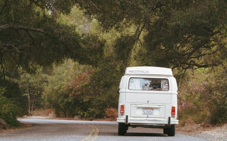 Biały klasyczny kamper typu van jadący wąską drogą przez gęsty, zielony las.
