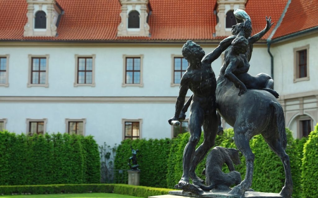 Brązowa rzeźba centaura z postaciami w Ogrodzie Waldsteńskim na tle białej barokowej kamienicy