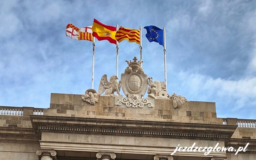 Cztery powiewające flagi Barcelony, Hiszpanii, Katalonii oraz Unii Europejskiej umieszczone nad kamienną balustradą z herbem.