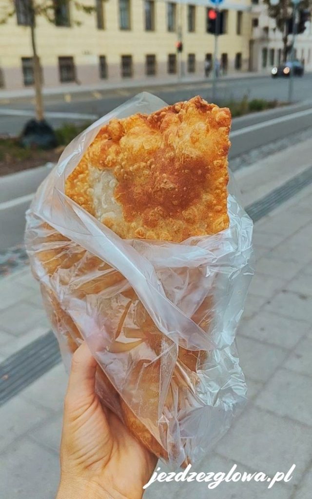 Dłoń trzymająca tradycyjną łotewską drożdżówkę (pirags) zawiniętą w papier na tle ulicy w Rydze