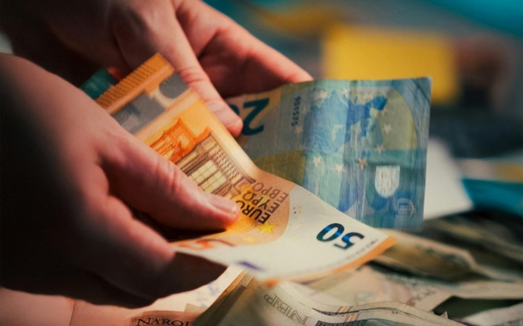 Dłonie osoby odliczającej banknoty o nominale 50 euro na tle rozmytego stosu pieniędzy.