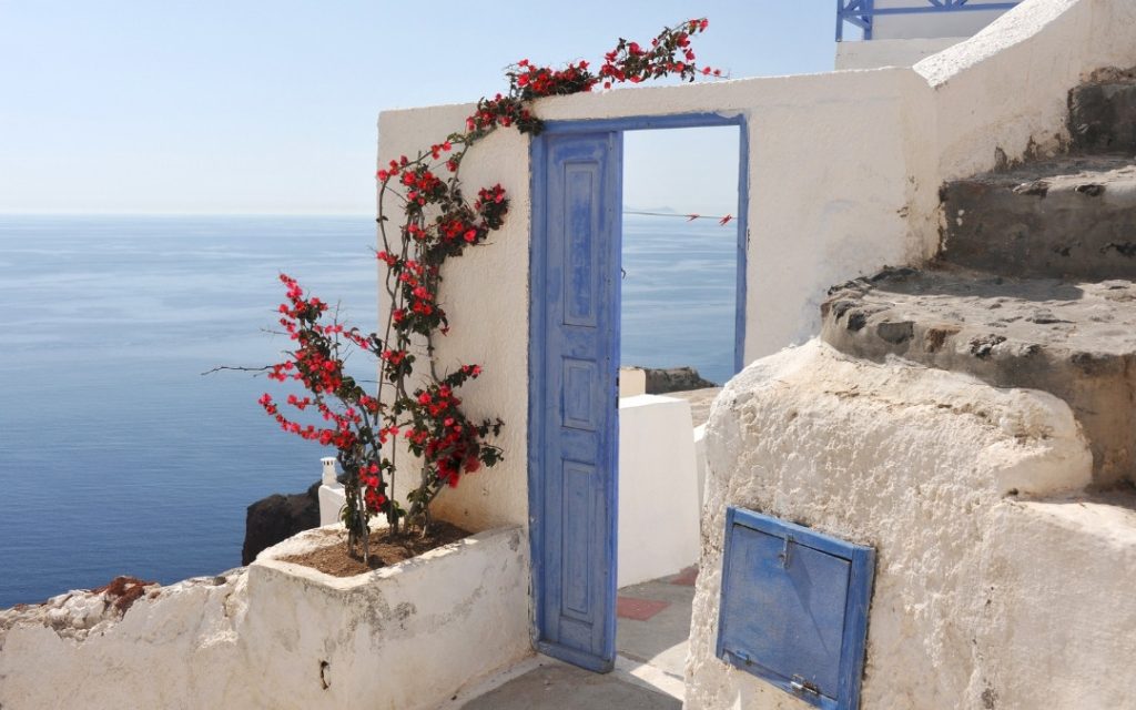 Fragment białej greckiej zabudowy z błękitną futryną drzwi i widokiem na morze.