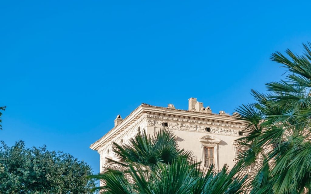 Górna część fasady Palazzo Barberini z bogatymi zdobieniami wśród palm na tle błękitnego nieba