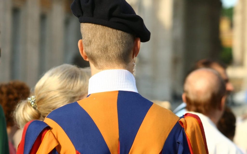Gwardzista Gwardii Szwajcarskiej widziany od tyłu w niebiesko-pomarańczowym mundurze i czarnym berecie wśród turystów