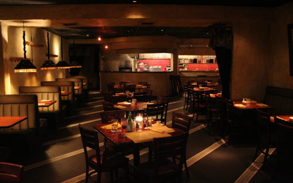Nastrojowe wnętrze włoskiej restauracji z ciemnymi meblami, nastrojowym oświetleniem i widocznym piecem do pizzy w tle