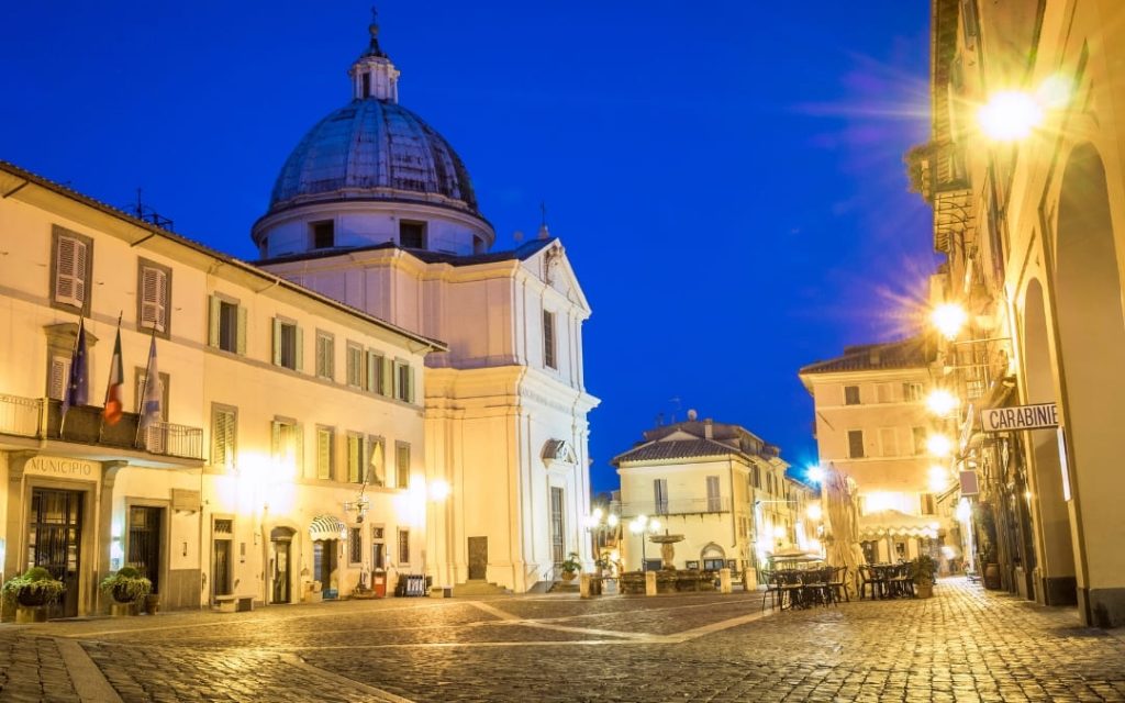 Oświetlony brukowany rynek w Castel Gandolfo z kościołem zwieńczonym kopułą i ratuszem w nocnej scenerii