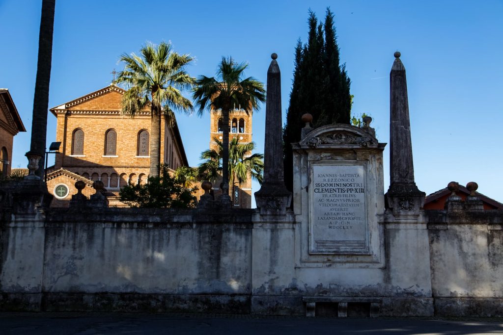 Zabytkowa bazylika otoczona palmami i kamiennymi obeliskami na Piazza dei Cavalieri di Malta w Rzymie