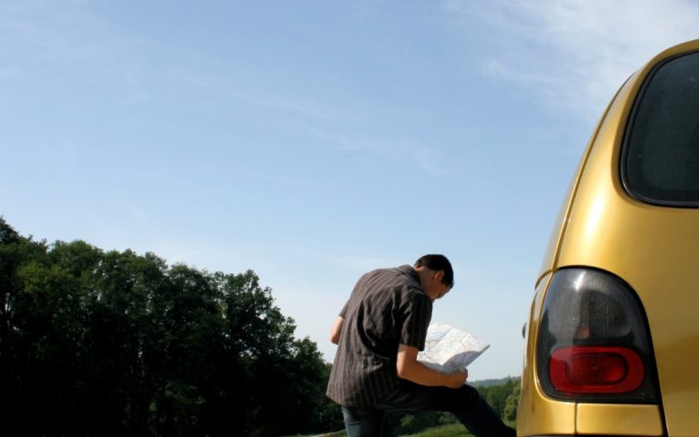 Podróżnik opierający się o żółty samochód i sprawdzający mapę drogową na tle błękitnego nieba i lasu.