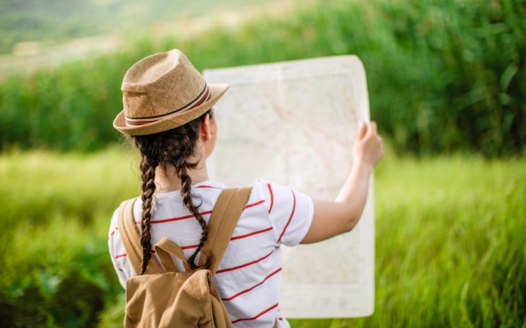 Podróżnik z plecakiem i w słomkowym kapeluszu sprawdzający mapę na tle zielonej łąki.