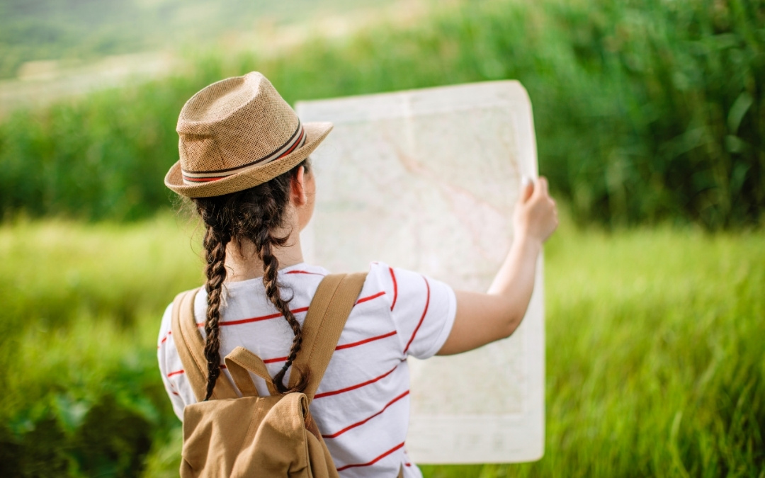 Podróżnik z plecakiem i w słomkowym kapeluszu sprawdzający mapę na tle zielonej łąki.