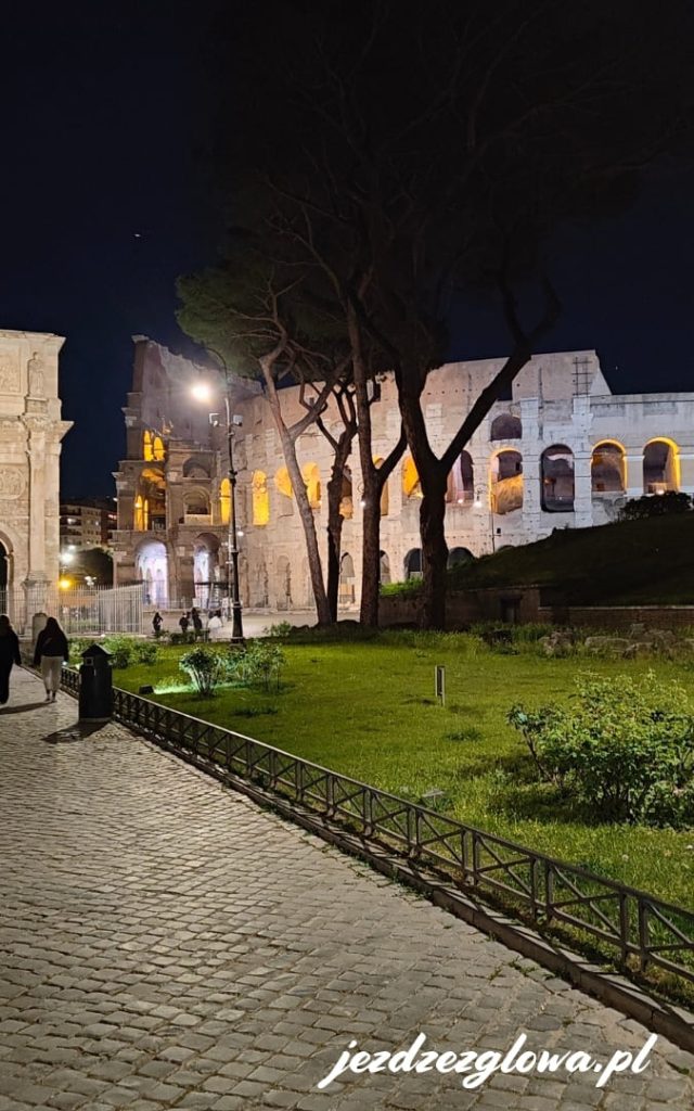 Podświetlone Koloseum nocą widoczne zza drzew przy brukowanej alejce w Rzymie