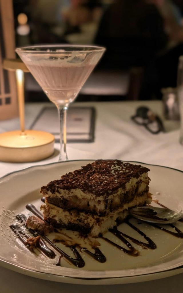 Porcja tiramisu z polewą czekoladową na talerzu i kieliszek koktajlu w tle