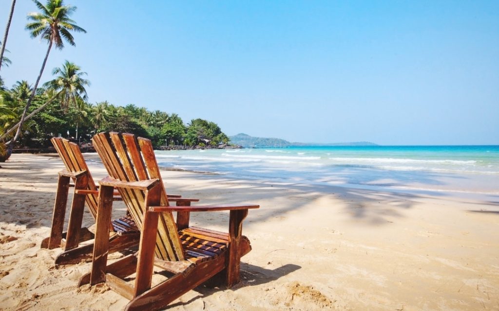 Puste drewniane fotele na białym piasku z widokiem na egzotyczną zatokę i palmy.