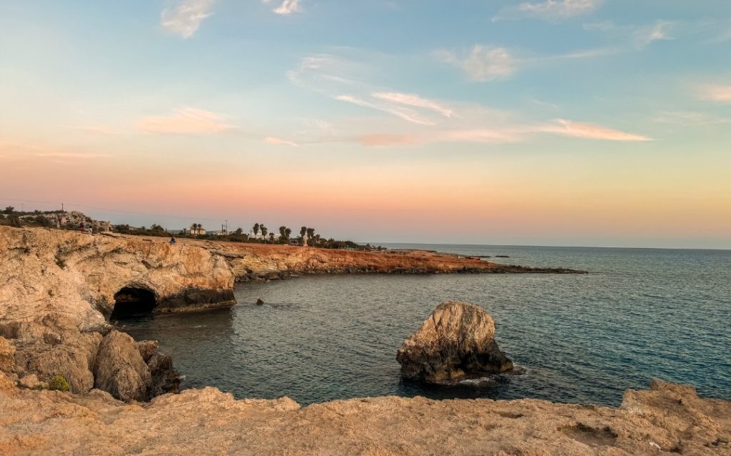 Skaliste wybrzeże Cypru z naturalnymi grotami i łukami skalnymi nad błękitnym morzem podczas złotego zachodu słońca.