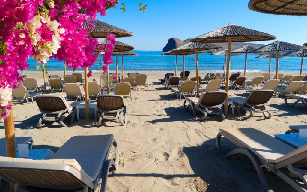 Słoneczna plaża na Krecie z parasolami ze słomy i różowymi kwiatami bugenwilli na pierwszym planie.
