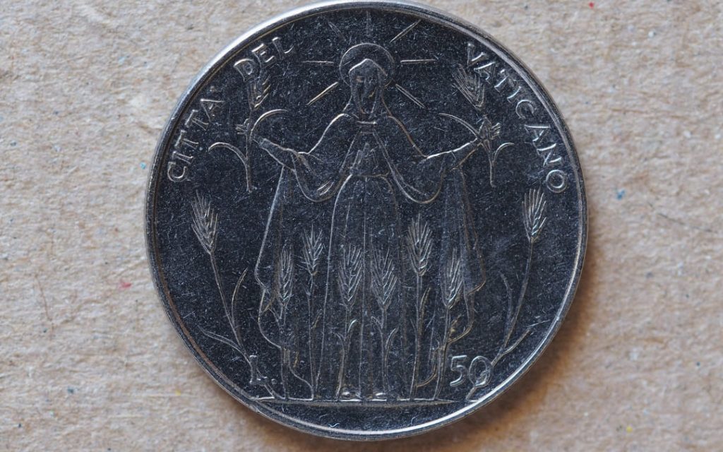 Srebrna moneta Città del Vaticano z rytą postacią świętej w aureoli wśród kłosów zboża, leżąca na jasnym tle