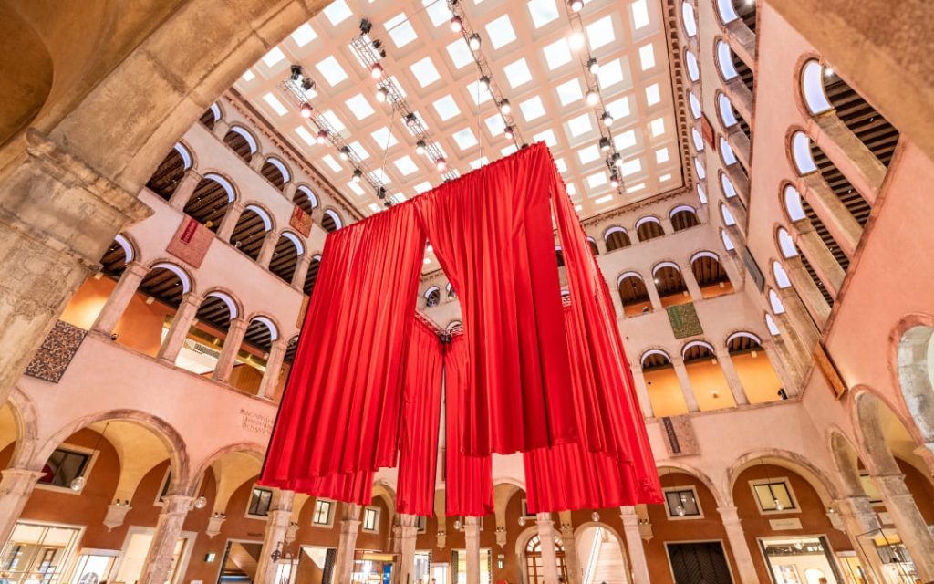 Ujęcie z dołu na przestrzenny dziedziniec Fondaco dei Tedeschi, czerwony baldachim i dzienne światło wpadające przez kwadratowe okna w suficie