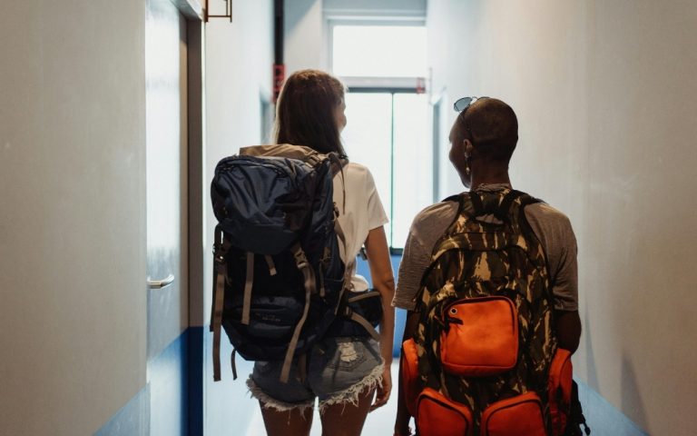 Widok od tyłu na dwie osoby z dużymi plecakami turystycznymi, idące hostelowym korytarzem w stronę jasnego okna.