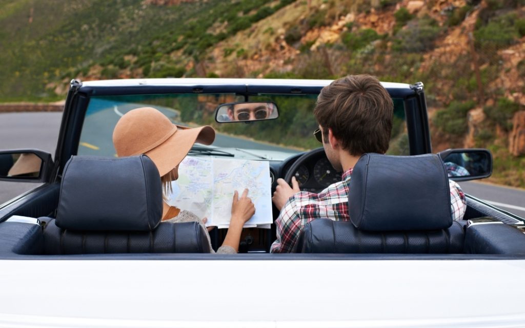 Widok z tyłu na parę jadącą białym kabrioletem, pasażerka w kapeluszu sprawdza trasę na papierowej mapie podczas podróży górską drogą.