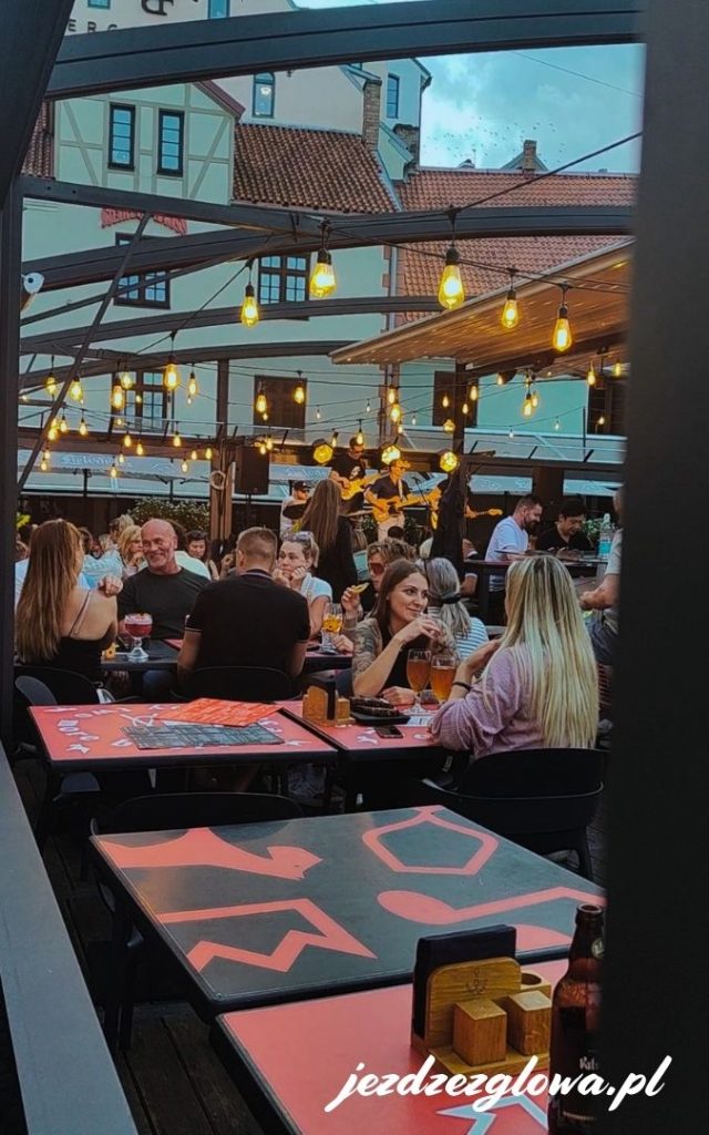 Wieczorny widok na pełen ludzi ogródek restauracyjny na Starym Mieście w Rydze, oświetlony girlandami żarówek