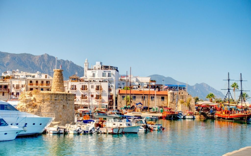 Zabytkowy port na Cyprze z zacumowanymi jachtami i łodziami rybackimi na tle gór i zabudowy.