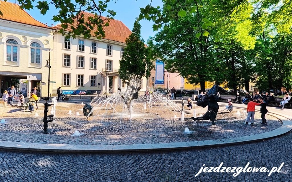 Fontanna Brunnen der Lebensfreude w Rostocku z rzeźbami postaci ludzi i zwierząt oraz ludźmi odpoczywającymi na ławeczkach w słoneczny dzień