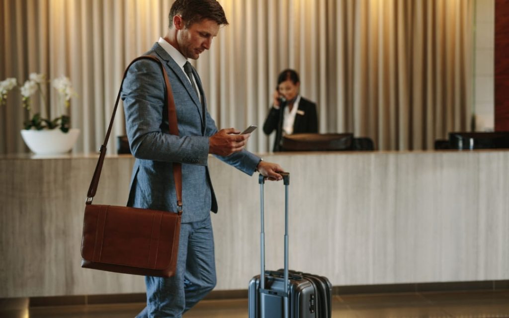 Mężczyzna w garniturze z walizką przeglądający telefon w hotelowym lobby przy recepcji