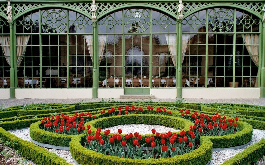 Oranżeria w ogrodach zamkowych z czerwonymi tulipanami i przystrzyżonymi żywopłotami, z widoczną za oknami salą z krzesłami