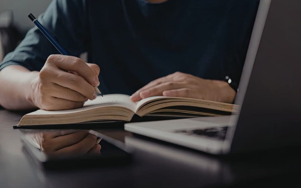 Osoba robiąca notatki ołówkiem w zeszycie przy otwartym laptopie na ciemnym biurku