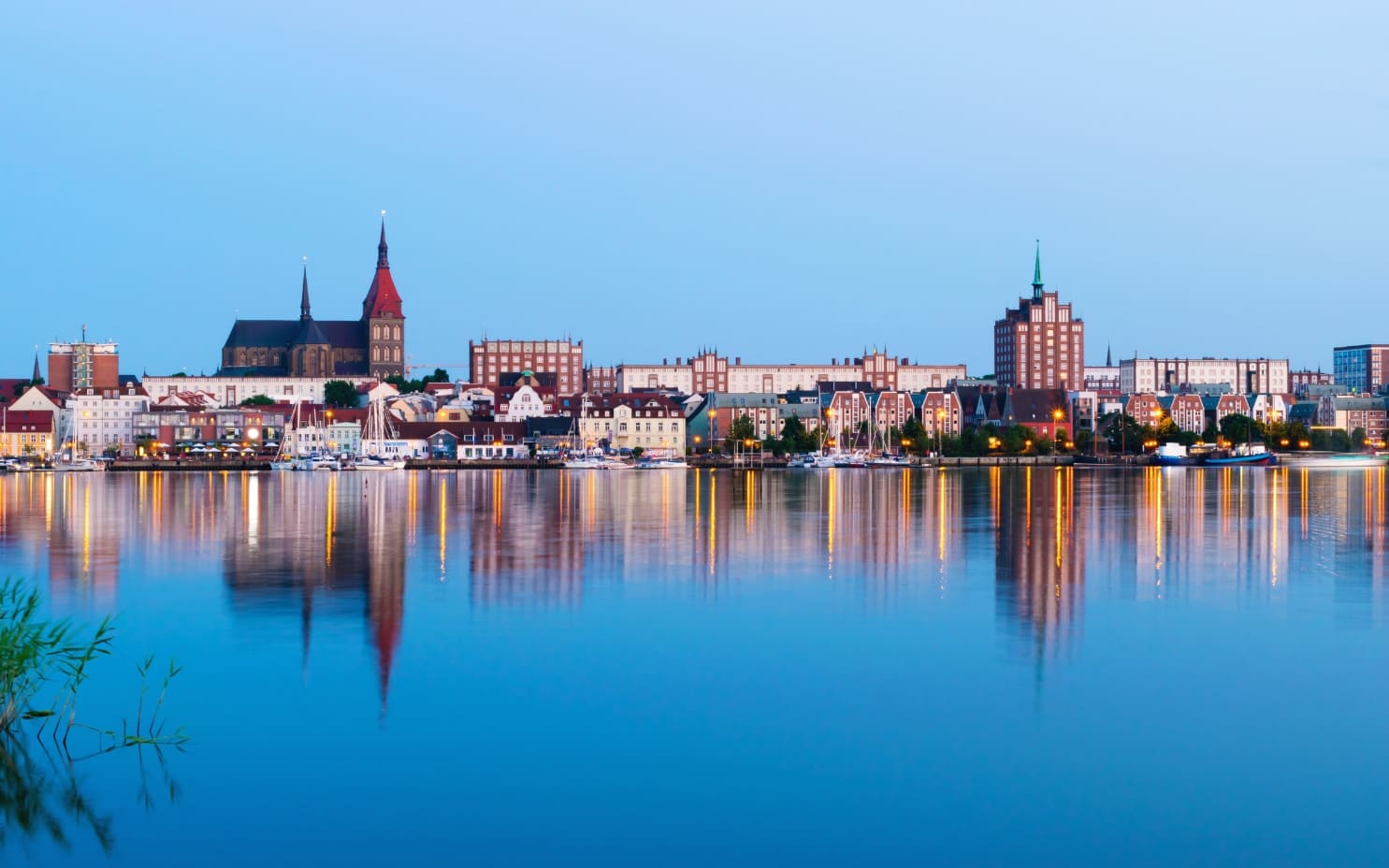 Port miejski Rostocku nad rzeką Warnow z odbijającą się w wodzie zabudową miasta i zacumowanymi jachtami