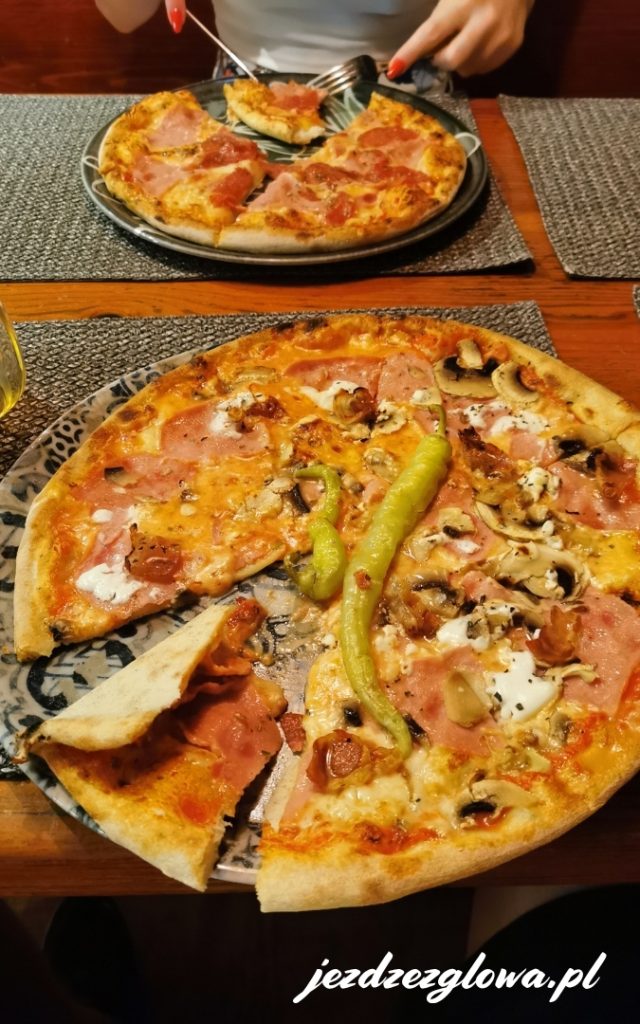 Pizza z szynką, pieczarkami i papryczką podana w restauracji, z drugim zamówieniem widocznym na stole w tle.
