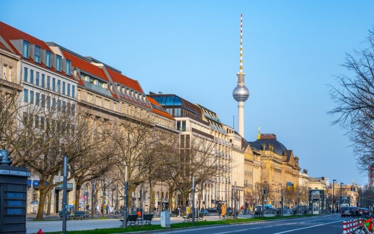 Aleja Unter den Linden w Berlinie z elegancką zabudową, ruchem ulicznym i wieżą telewizyjną Fernsehturm widoczną nad dachami budynków
