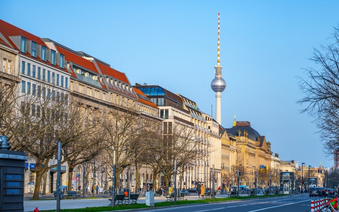 Aleja Unter den Linden w Berlinie z elegancką zabudową, ruchem ulicznym i wieżą telewizyjną Fernsehturm widoczną nad dachami budynków