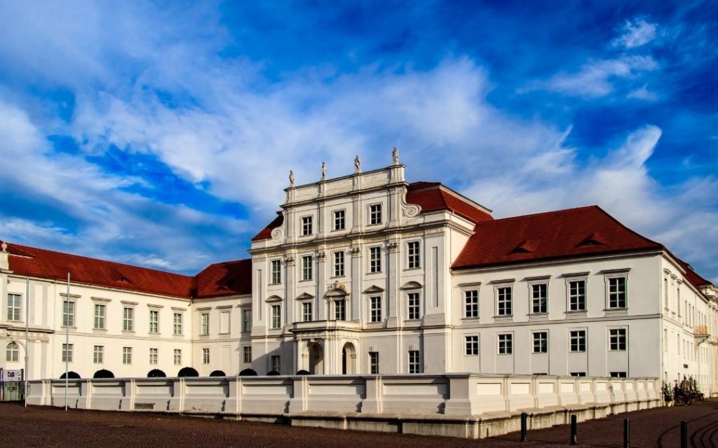Barokowy pałac Oranienburg z jasną fasadą, czerwonym dachem i ozdobnym frontonem, sfotografowany pod błękitnym niebem z chmurami