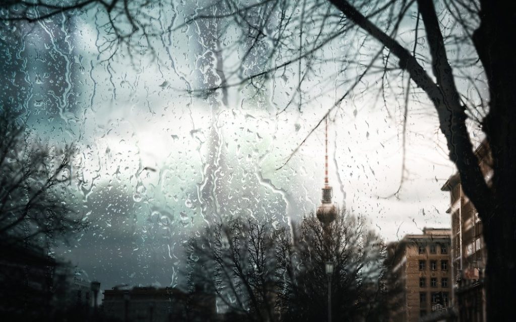 Deszczowy widok na Berlin przez szybę pokrytą kroplami wody, z wieżą Fernsehturm widoczną w oddali między drzewami i miejską zabudową