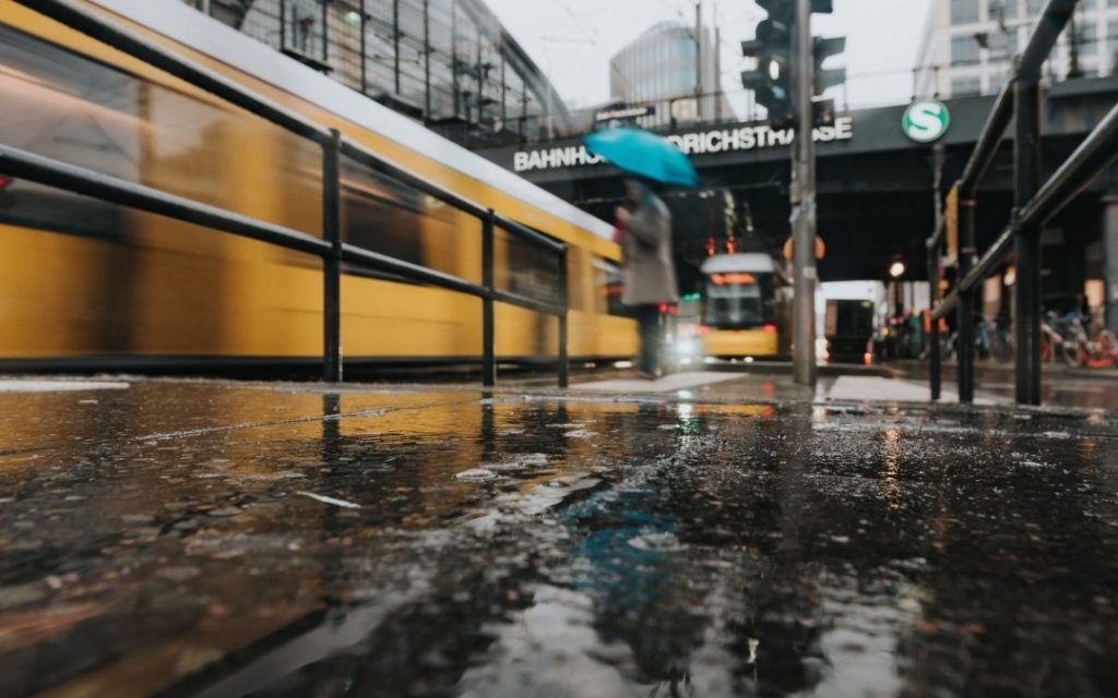 Deszczowy widok przy stacji Bahnhof Friedrichstraße w Berlinie, z rozmytym żółtym tramwajem, mokrym chodnikiem i odbiciami świateł w kałużach