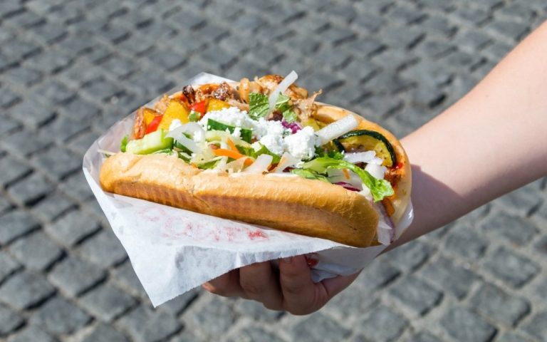 Döner kebab trzymany w dłoni na brukowanej ulicy, wypełniony mięsem, grillowanymi warzywami, świeżą sałatą, cebulą i białym serem