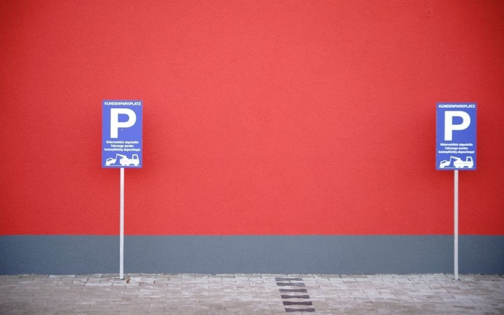 Dwa niebieskie znaki parkingu dla klientów ustawione przed intensywnie czerwoną ścianą z szarym pasem u dołu