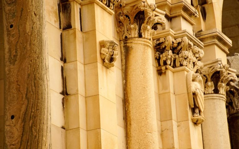 Fragment zabytkowej kamiennej fasady z ozdobnymi kolumnami, bogato rzeźbionymi kapitelami oraz dekoracyjnymi figurami umieszczonymi na elewacji budynku