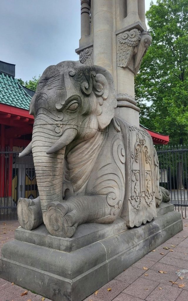 Kamienna rzeźba słonia przy Bramie Słoni Elefantentor w Zoo Berlin, z ozdobną kolumną i zielenią drzew w tle