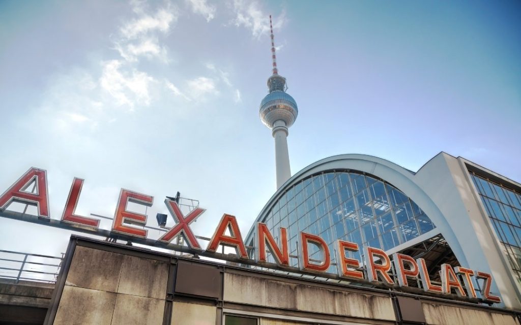 Napis Alexanderplatz na dachu stacji w Berlinie, z wieżą telewizyjną Fernsehturm i przeszkloną halą dworcową w tle
