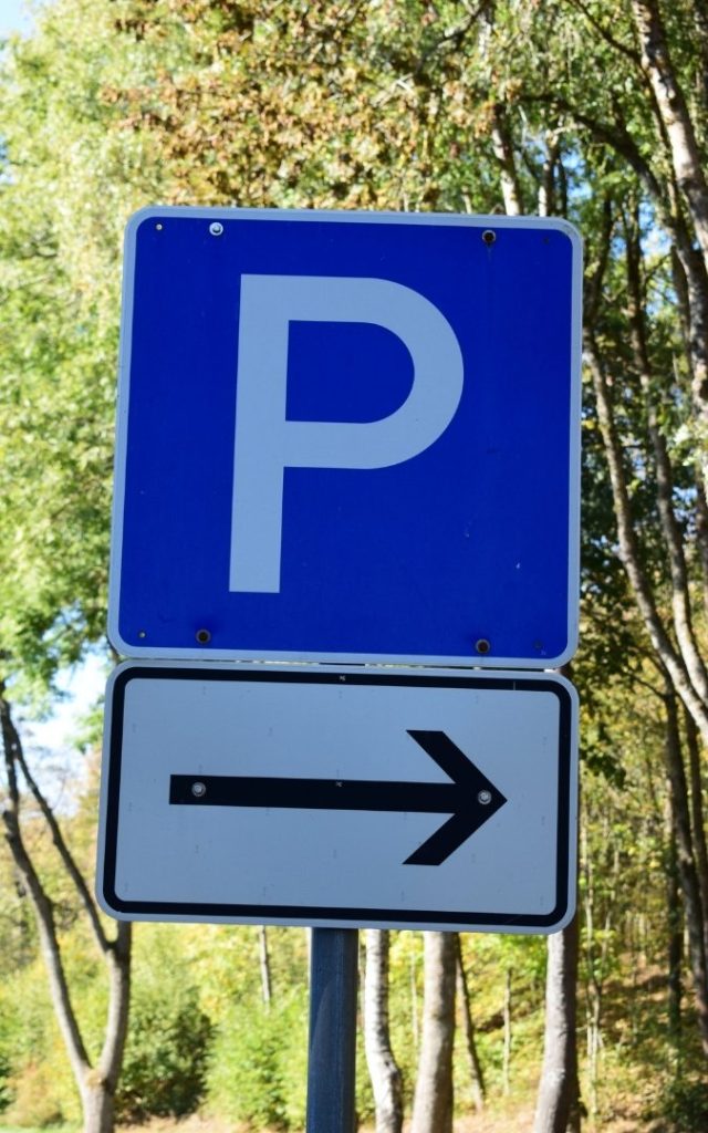 Niebieski znak parkingu z białą literą P i strzałką kierunkową w prawo, ustawiony przy drodze na tle zielonych drzew