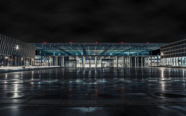 Nocny widok na terminal lotniska Berlin Brandenburg BER, z podświetloną szklaną fasadą, mokrą nawierzchnią placu i odbiciami świateł
