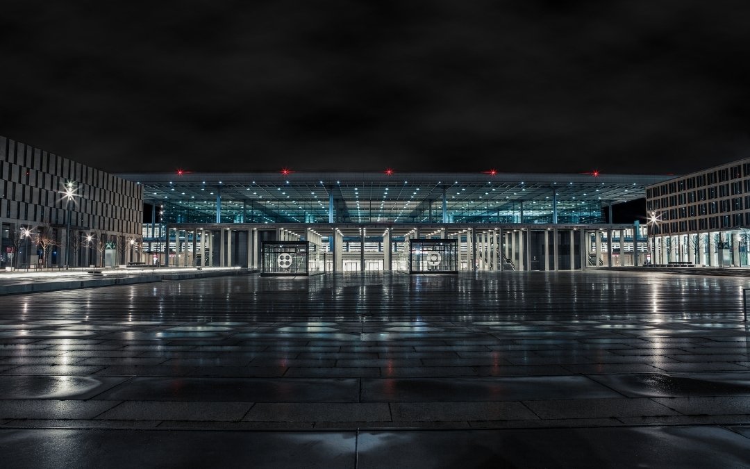 Nocny widok na terminal lotniska Berlin Brandenburg BER, z podświetloną szklaną fasadą, mokrą nawierzchnią placu i odbiciami świateł