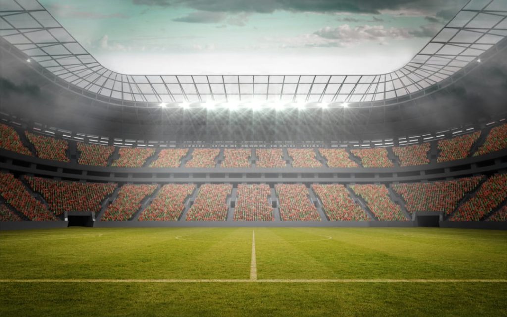Nowoczesny stadion piłkarski z oświetloną murawą, zadaszonymi trybunami wypełnionymi kibicami oraz reflektorami nad boiskiem