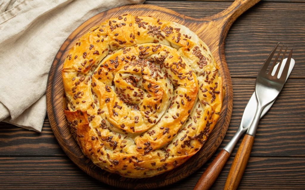 Okrągły burek z chrupiącego ciasta filo posypany ziarnami lnu, podany na drewnianej desce
