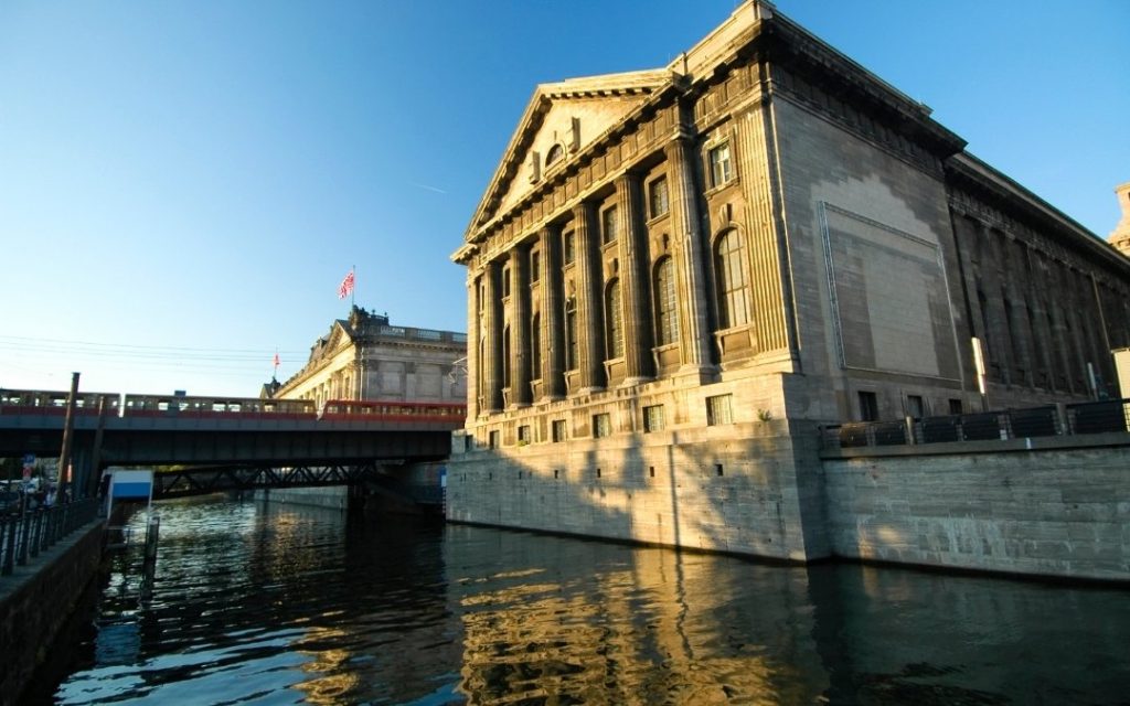 Pergamonmuseum w Berlinie widziane od strony kanału, z klasycystyczną fasadą oświetloną słońcem, odbiciem w wodzie i przejeżdżającym pociągiem w tle