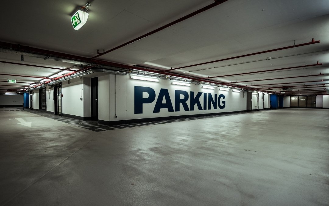 Podziemny parking z dużym napisem Parking na ścianie, pustą betonową przestrzenią, oświetleniem sufitowym i oznaczeniami ewakuacyjnymi