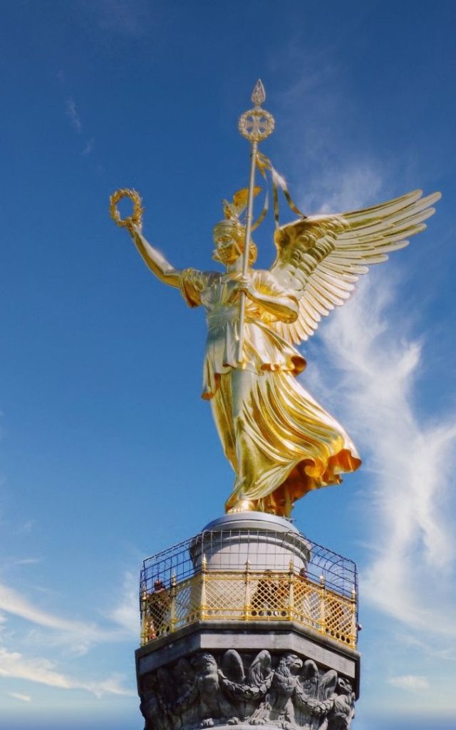 Pozłacana figura Wiktorii na szczycie berlińskiej Siegessäule, ukazana z bliska z wieńcem laurowym, skrzydłami i błękitnym niebem w tle
